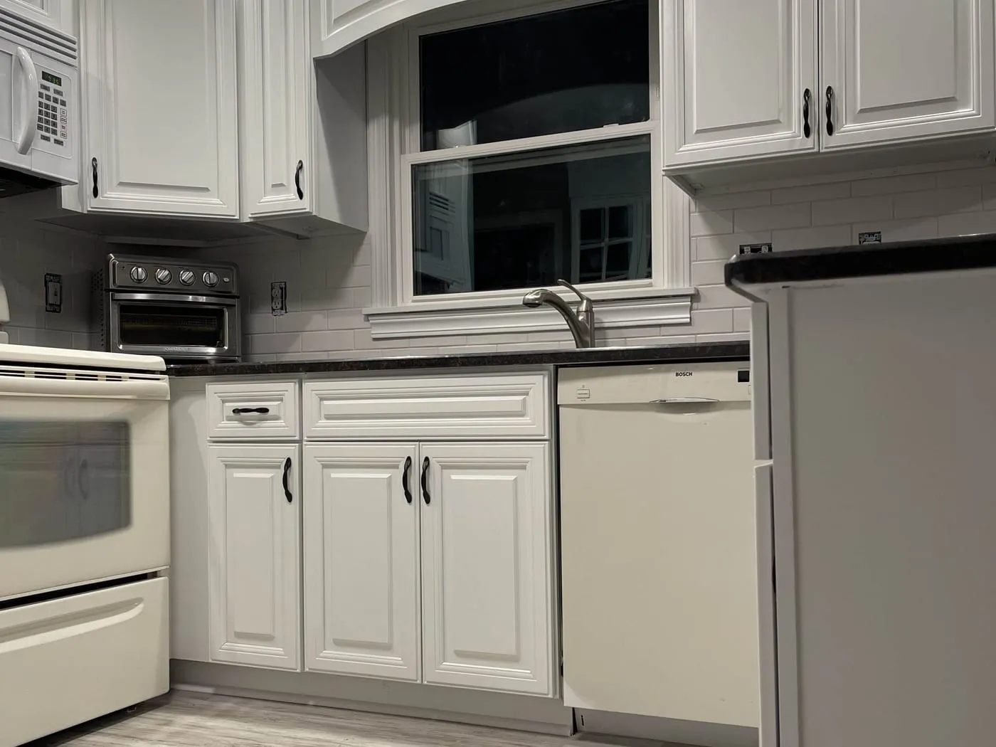 The same kitchen finished, with white painted cabinets, a new subway tile backsplash, dark counters and new flooring