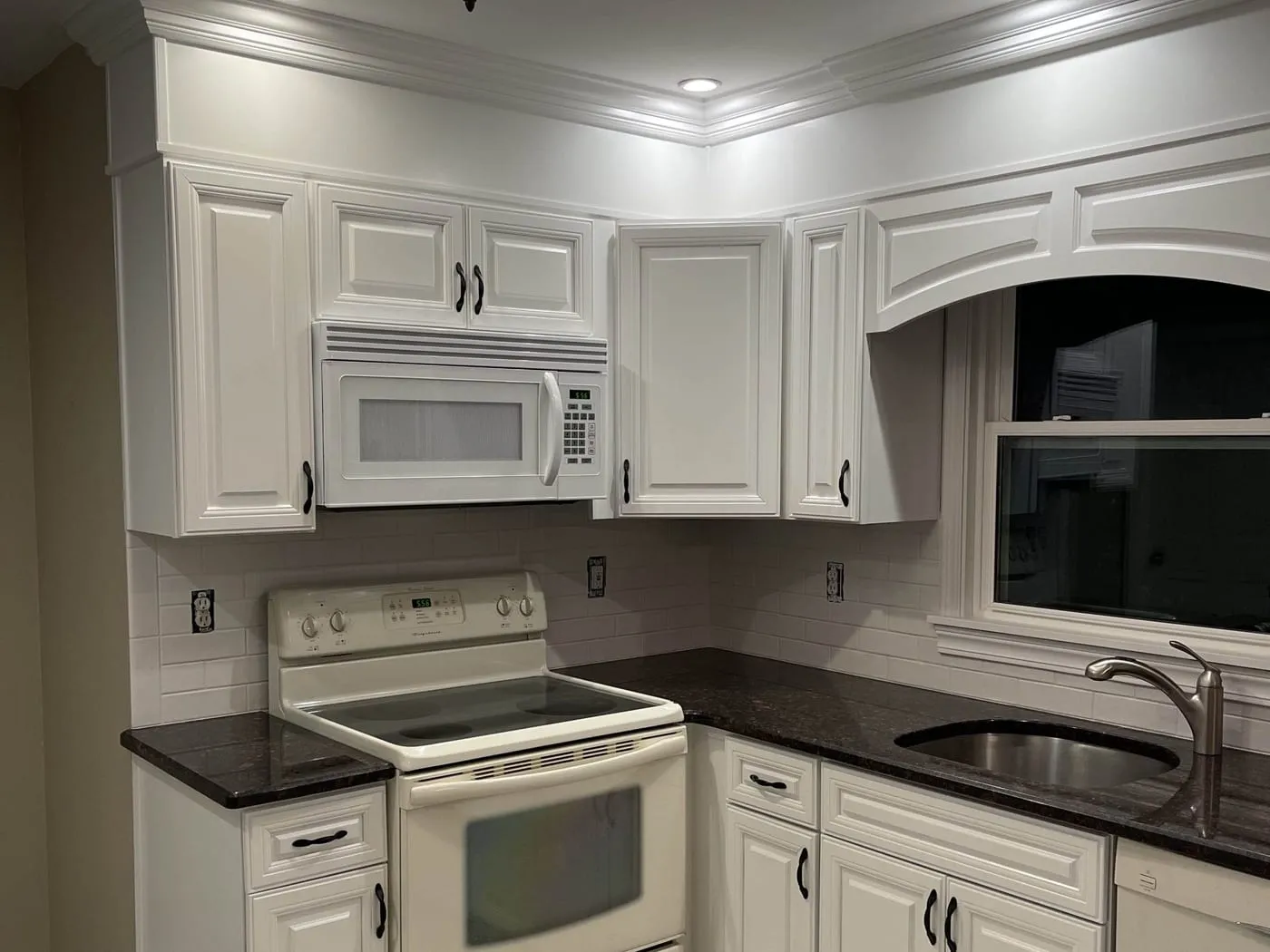 The same kitchen after cabinet painting, with smooth white cabinets, a new subway tile backsplash and dark counters