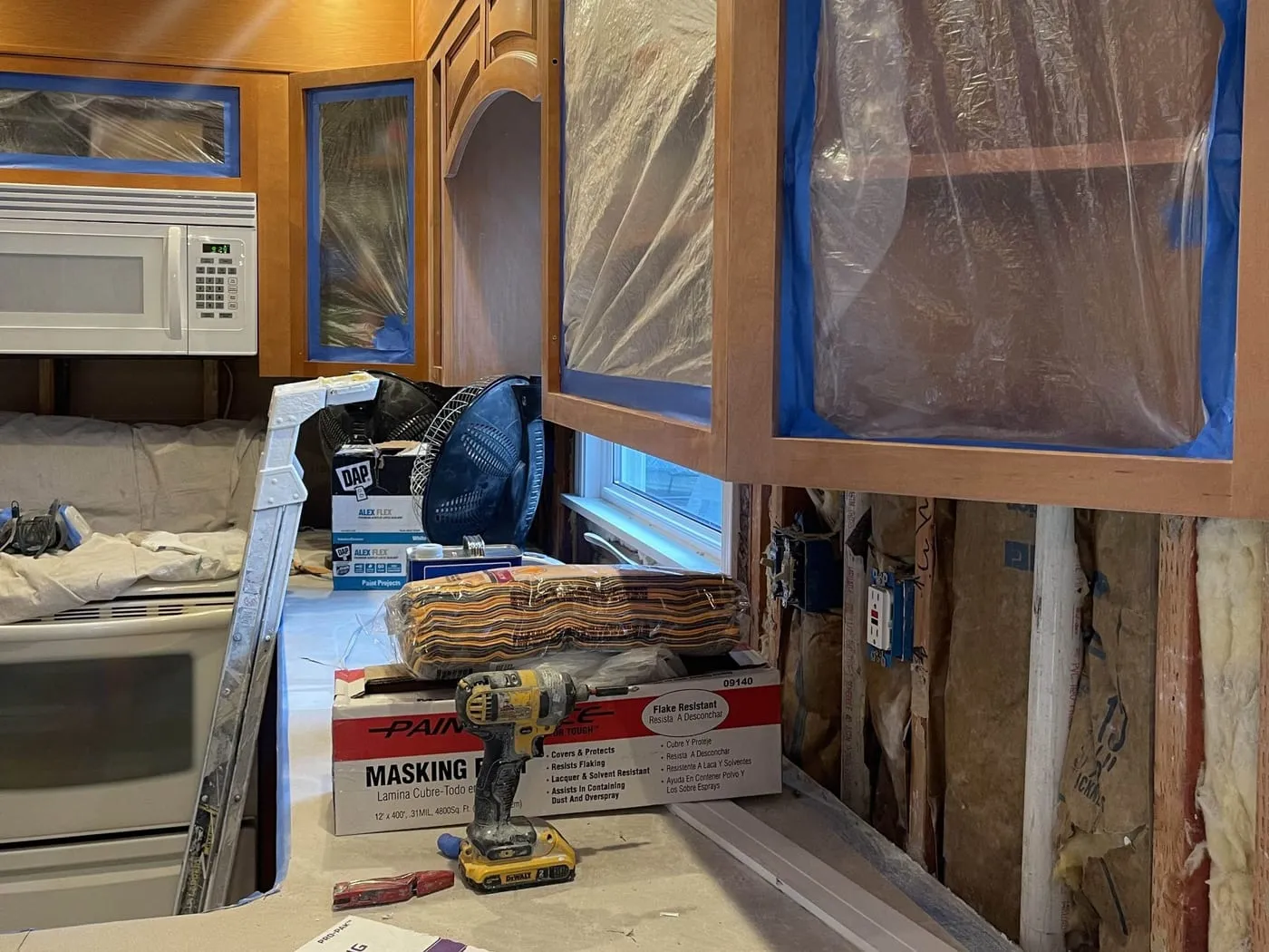 Kitchen mid-remodel with the old backsplash and drywall removed down to the studs and the cabinets masked for painting