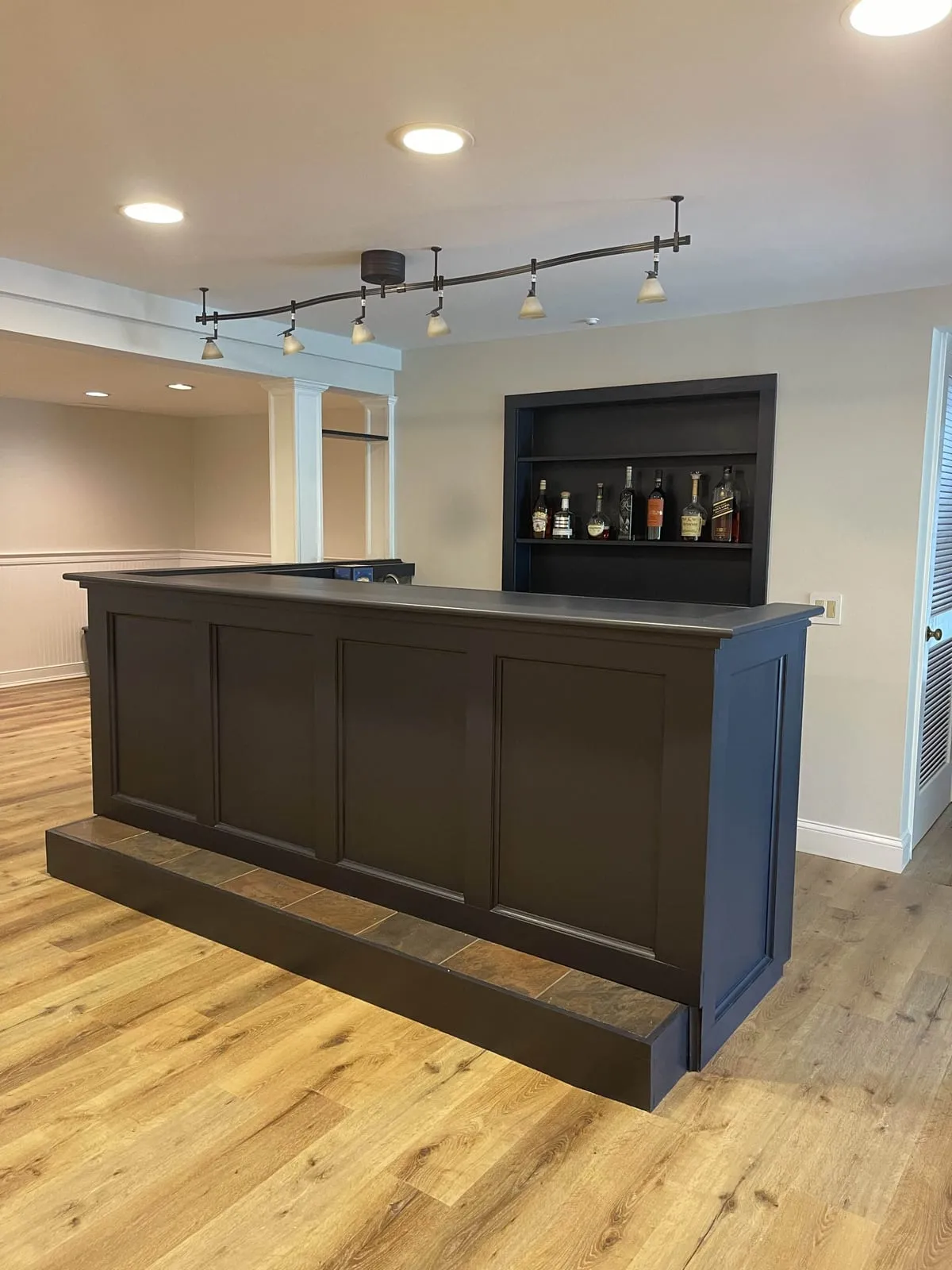 Finished lower level with a dark painted home bar on a tiled step and built-in shelving on the wall behind it