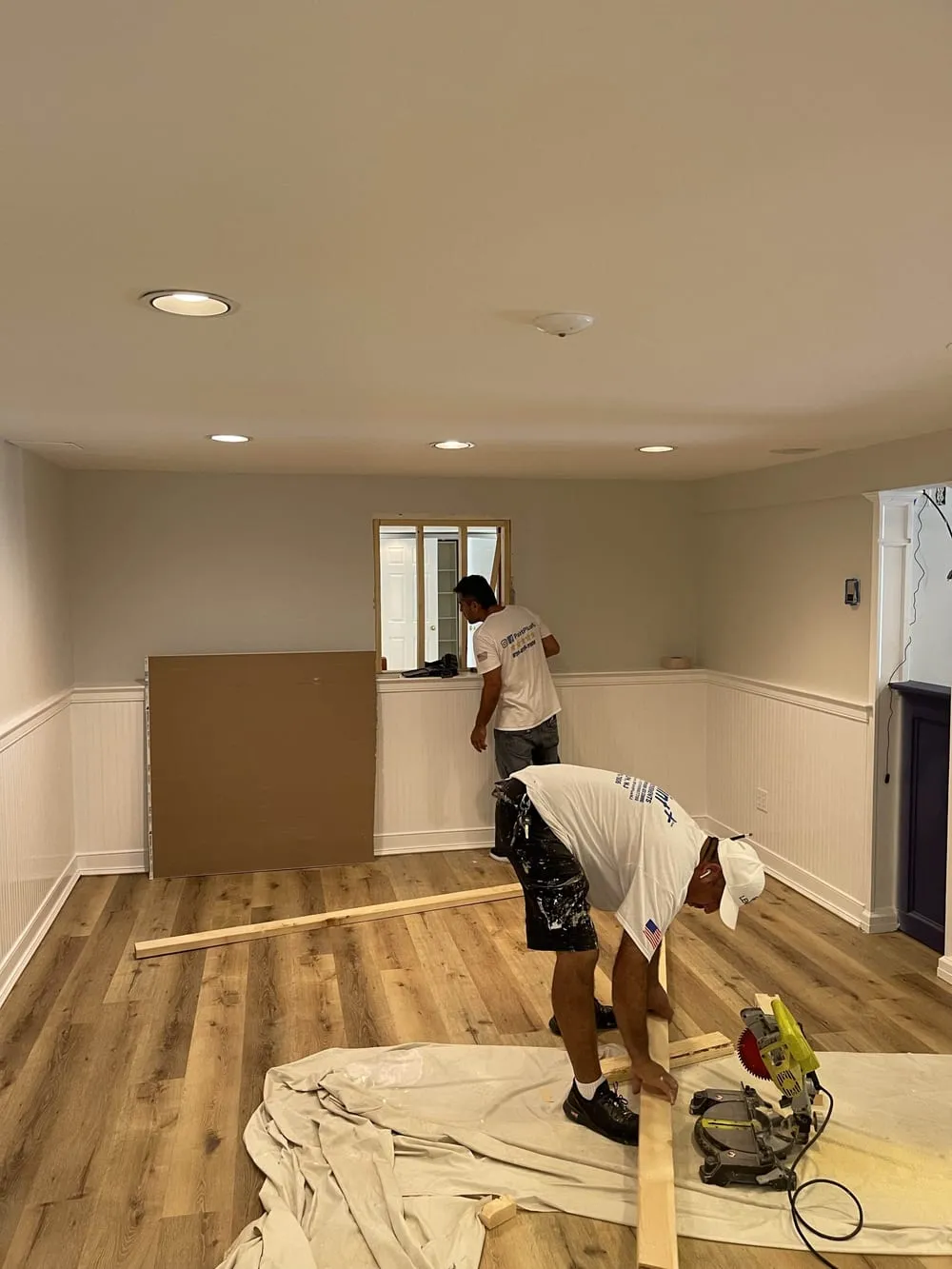 Paint Plus crew cutting and fitting trim in a lower-level room with wainscoting and plank flooring