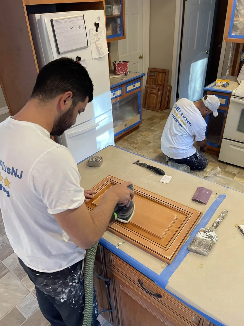 Paint Plus crew sanding cabinet doors on a protected counter while another member preps the lower cabinets