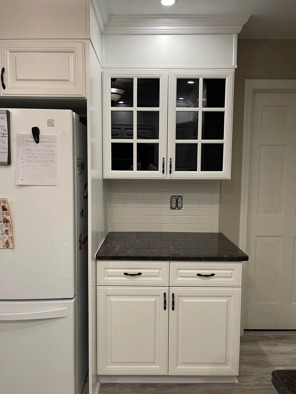 Glass-front cabinet and counter nook painted white, with dark granite and a subway tile backsplash