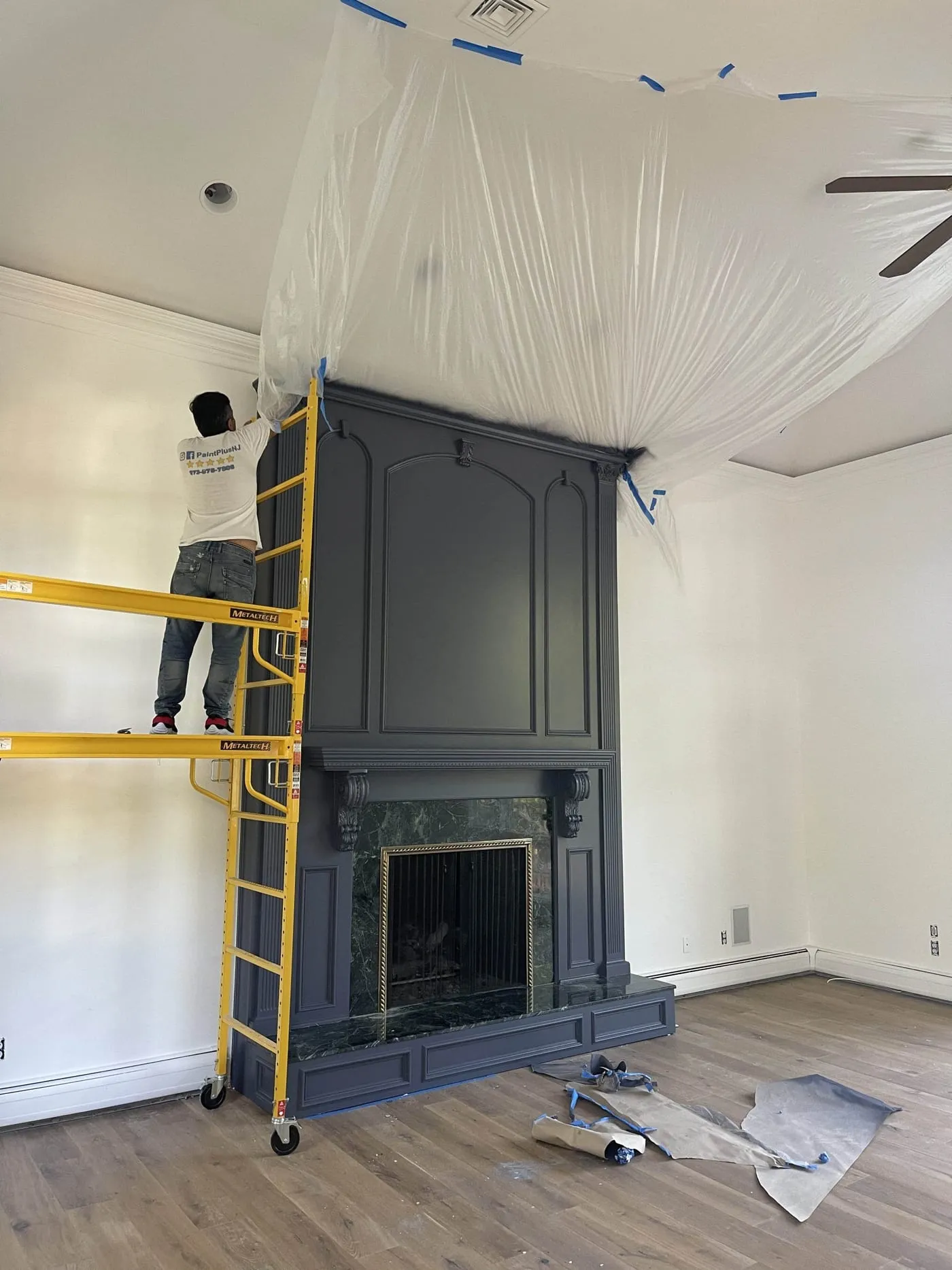 Paint Plus painter on scaffolding painting a fireplace surround navy, with the ceiling sealed off in plastic