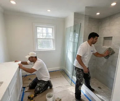 Bathroom remodel in progress, with a crew member fitting a white vanity and another cleaning grout on a gray tile shower