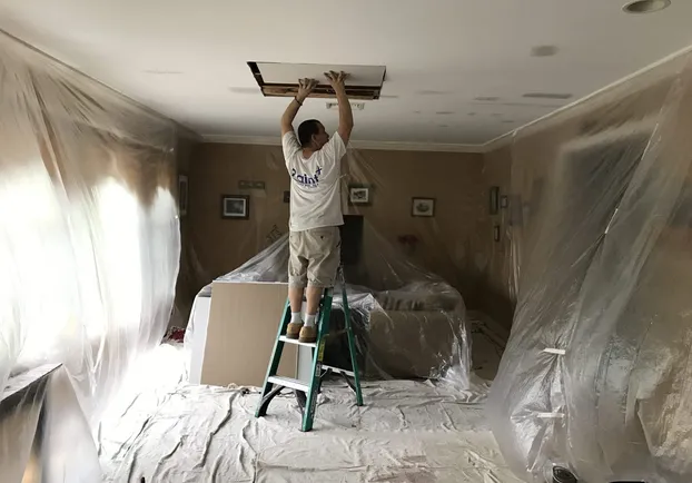 Room sealed floor to ceiling in plastic sheeting while a Paint Plus painter works on the ceiling from a stepladder